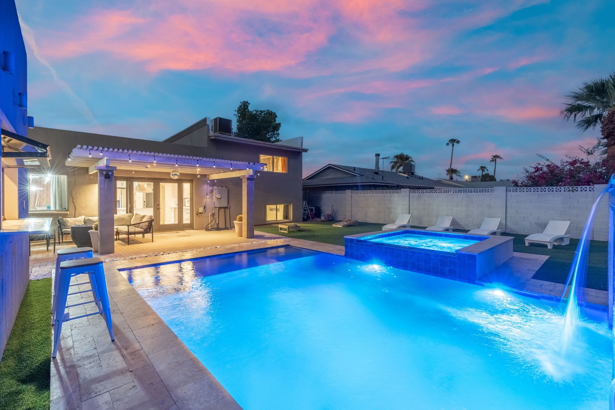 A swimming pool with a hot tub and lounge chairs, a covered patio area, and a view of the sky at dusk.