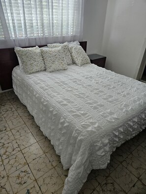 A neatly made bed with a white quilt and pillows, a bedside table, and a window with sheer curtains. - Spacious home available for Day of the Dead! (Oaxaca de Juárez)
