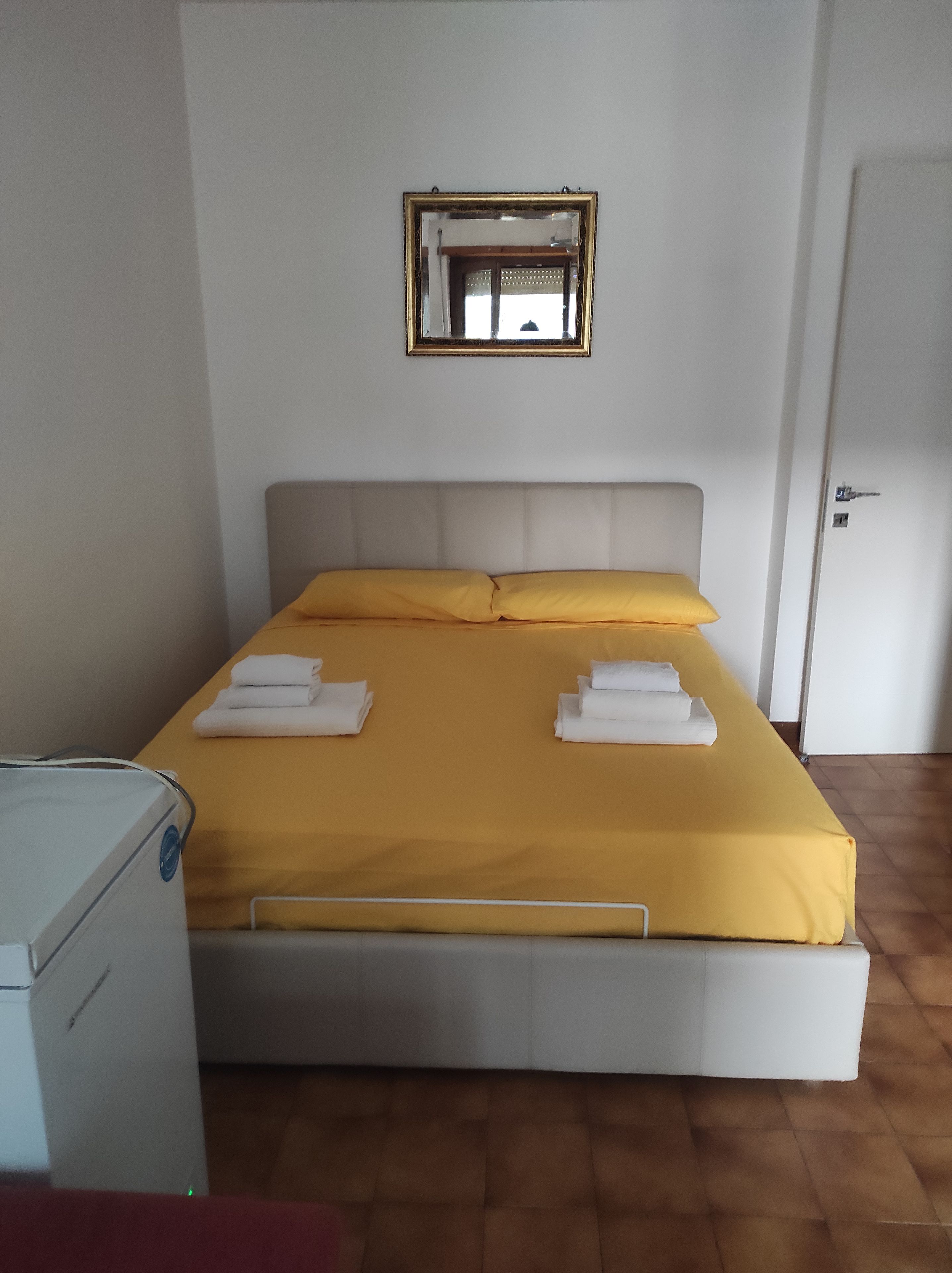 A single-bed room with a yellow bedspread, a bedside table with a lamp, and a mirror on the wall.
