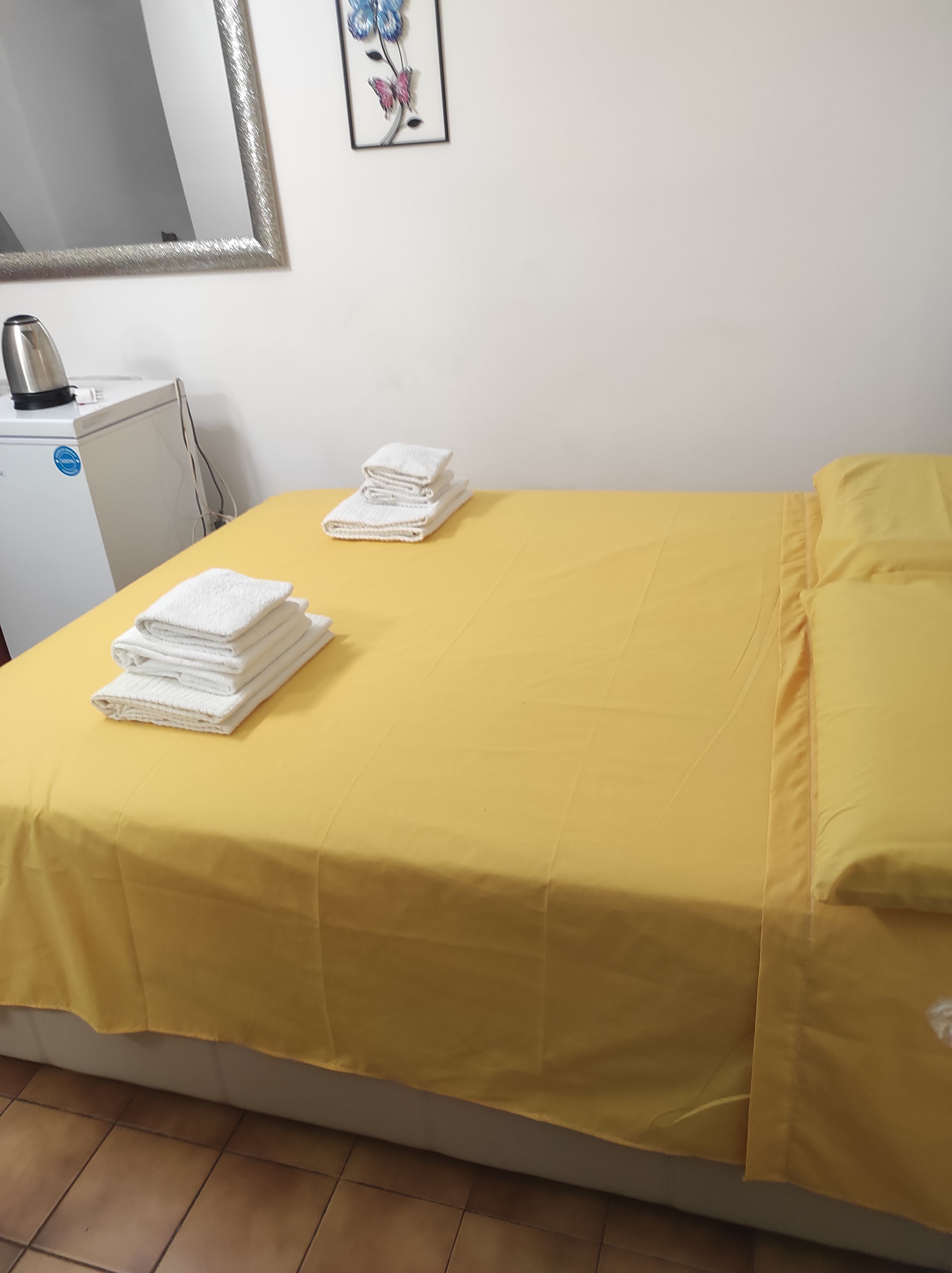A room with a bed covered in yellow bedding, a stack of white towels on the bed, a white radiator, and a kettle on a small white appliance.