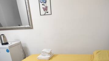 A room with a bed covered in yellow bedding, a stack of white towels on the bed, a white radiator, and a kettle on a small white appliance.