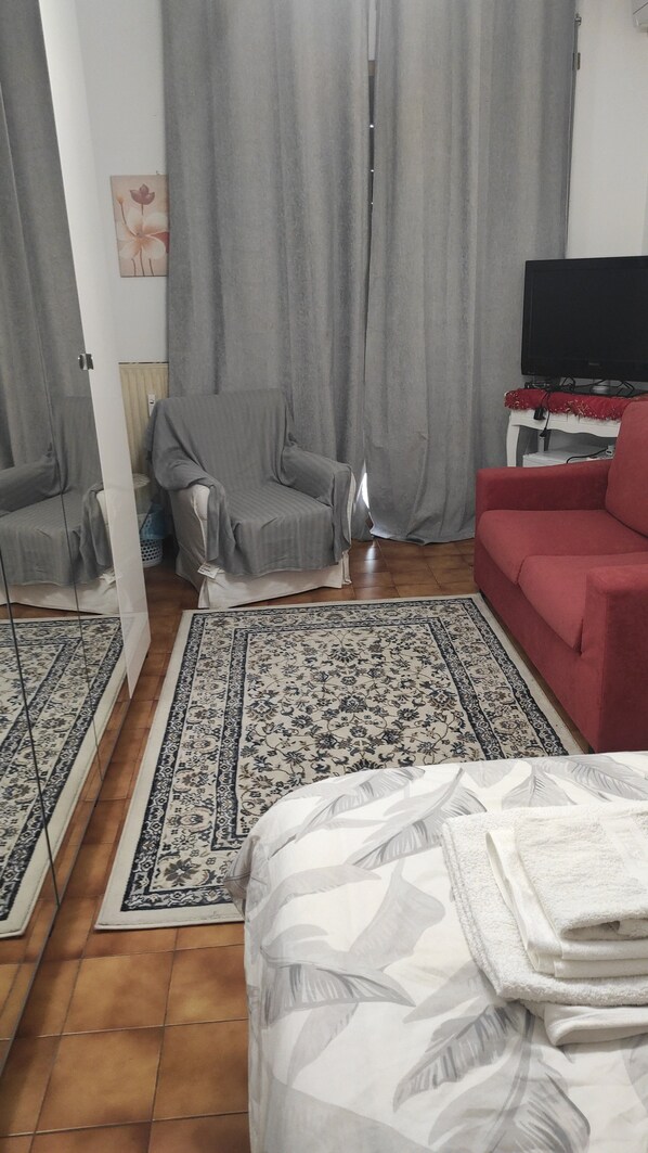 A room with a bed, a red sofa, a television, and a patterned rug. - New Listing (Roma)