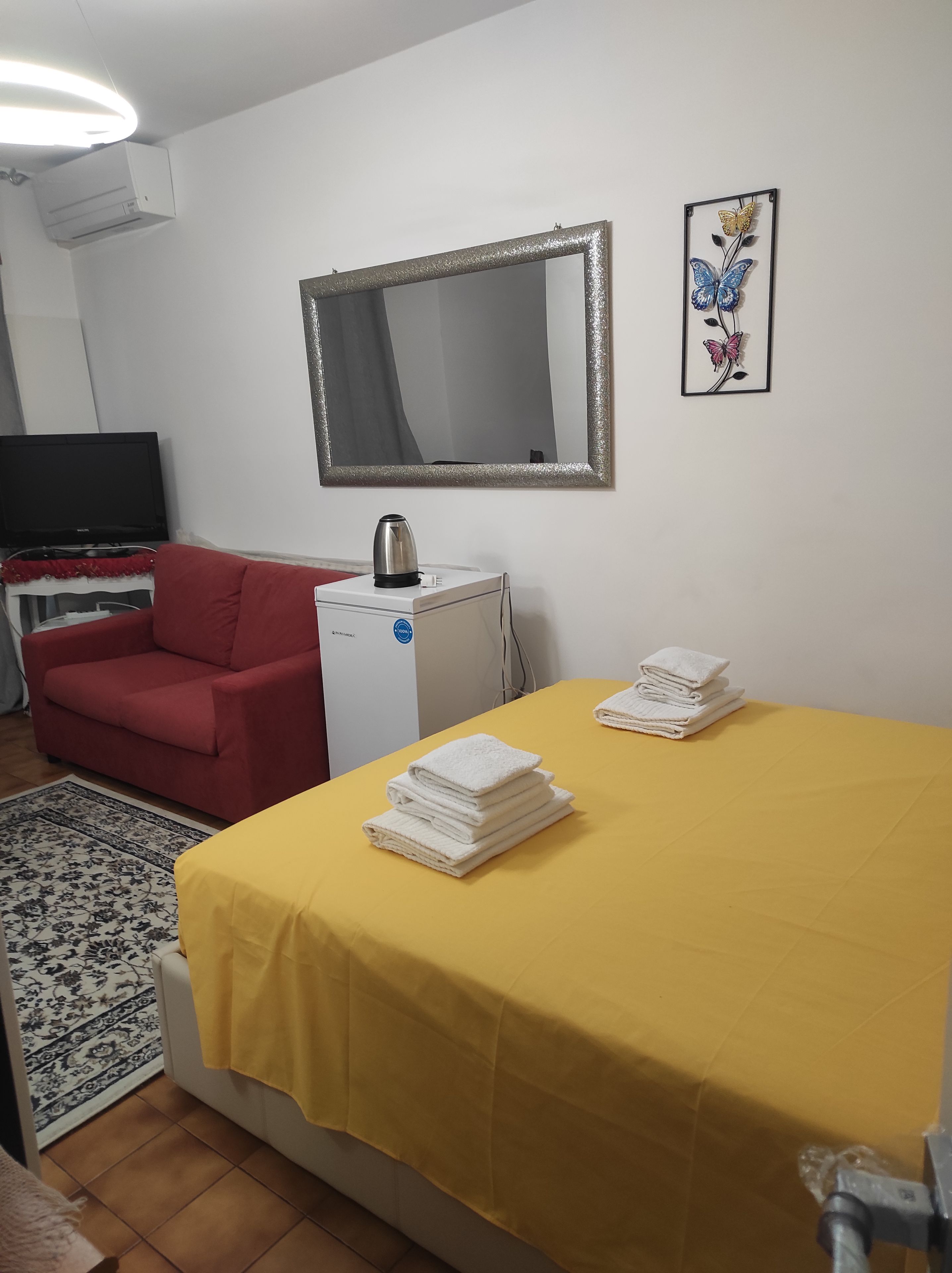A room with a bed, a red chair, a small table with a kettle, a flat-screen TV, and a framed artwork on the wall.
