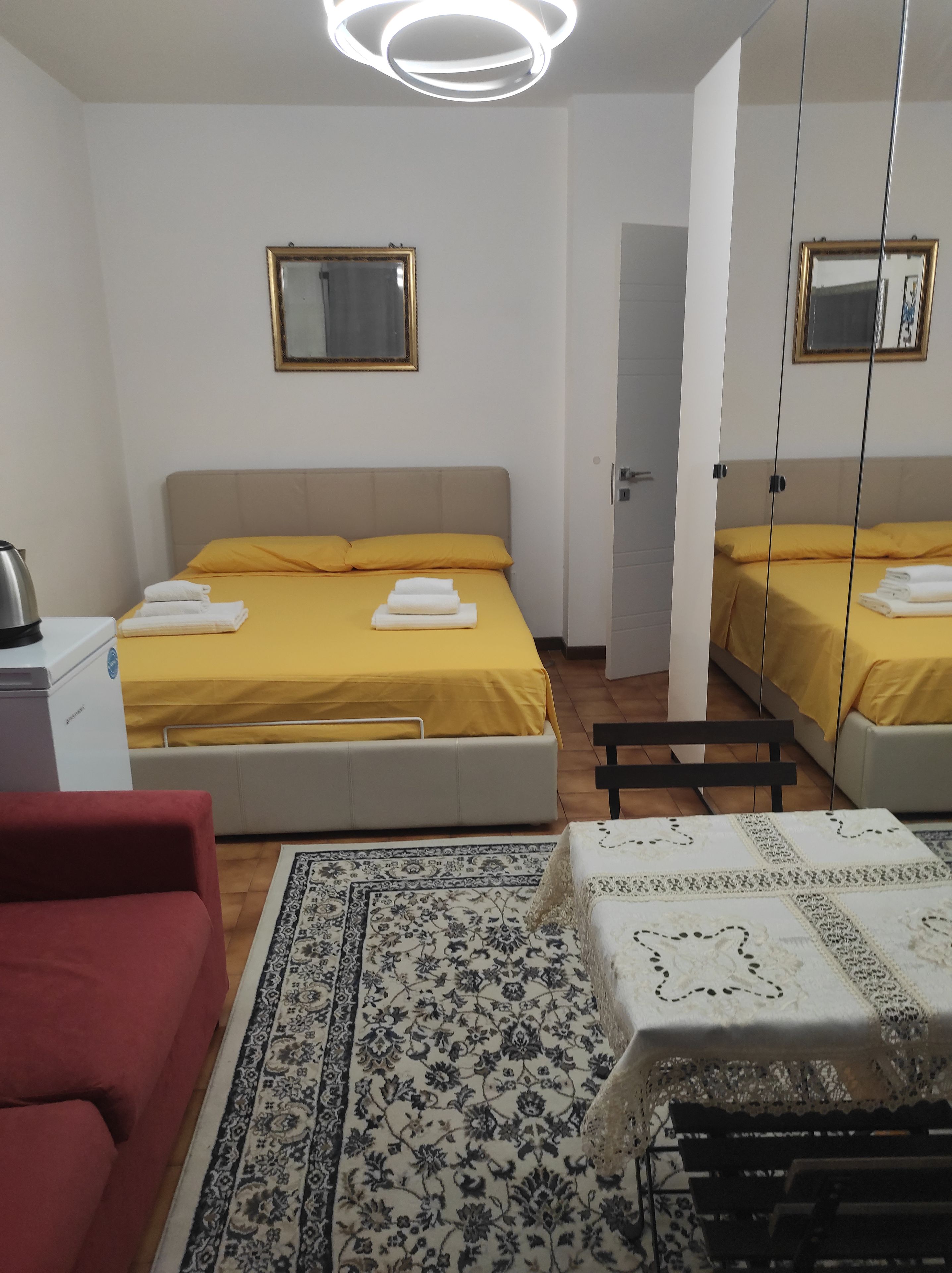 A hotel room with a bed, a desk, a chair, a red sofa, a patterned rug, and a mirror.