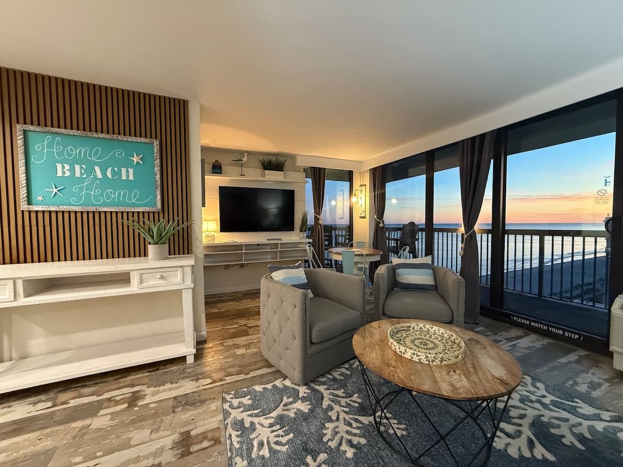A modern living room with a large TV, comfortable seating, a view of the beach, and a sign that says 'Home Beach Home'.
