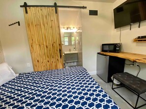 A bedroom with a bed, a desk, a television, and a bathroom visible through a sliding door.