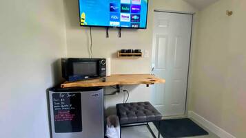 A compact living space with a flat-screen TV mounted on the wall, a microwave, a mini-fridge, a wooden shelf, and a small seating area.