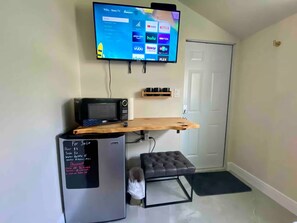 Un espacio habitable compacto con un televisor de pantalla plana en la pared, un microondas, un refrigerador pequeño, un estante de madera y una pequeña zona de estar.