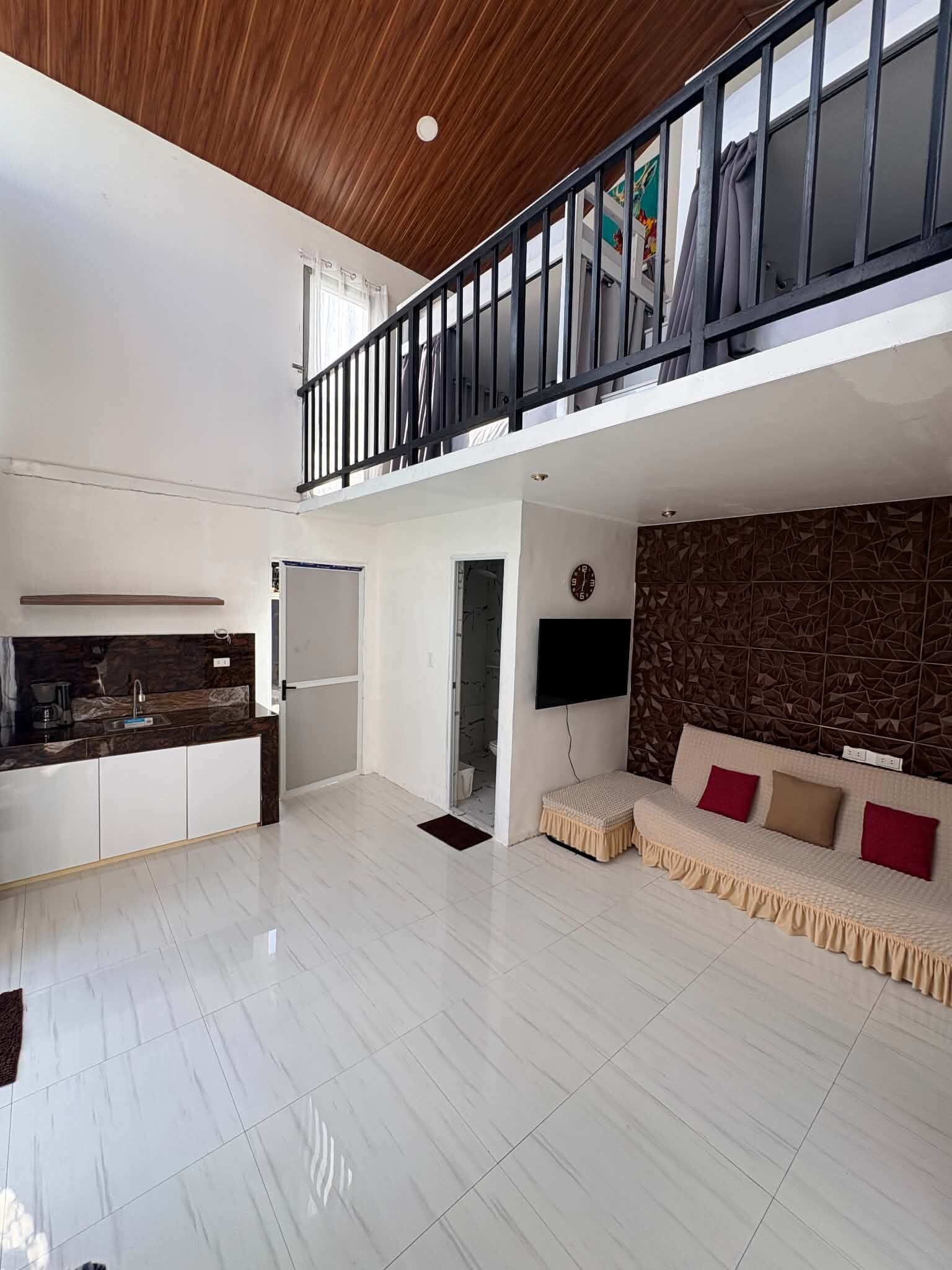 A spacious living area with a high ceiling, a balcony with a railing, a flat-screen TV mounted on the wall, and a sofa with cushions.