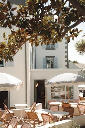 An outdoor seating area with wicker chairs and tables, umbrellas, and a building with windows and doors in the background. - HOTEL SAINT-ENOGAT (Dinard)