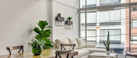 A modern living room with a dining table, sofa, and chairs, large windows, and potted plants.