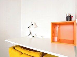 Ein weißer Schreibtisch mit einer weißen Schreibtischlampe, ein gelber Stuhl und eine orangefarbene Aufbewahrungsbox mit rosa Innenraum.