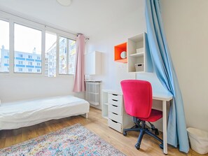 Ein Schlafzimmer mit Bett, Schreibtisch, Stuhl, Heizkörper, Fenster und Blick auf Gebäude.