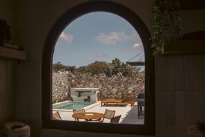Vista a través de un ventanal de arco que muestra un área de piscina con pared de piedra, sillas de descanso y una bañera de hidromasaje.