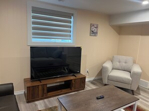 A living room with a flat-screen TV on a wooden stand, a grey armchair, and a coffee table. - Mayland Heights Retreat (Calgary)