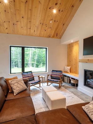 A cozy living room with a brown leather sofa, two rocking chairs with patterned cushions, a small round table, and a bench with a pillow. A fireplace is visible on the right, and there is a large window with a view of trees. - NEW Cozy home located within minutes to Sugarloaf (Carrabassett Valley)
