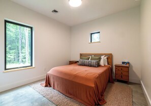 A bedroom with a large window, a bed with a wooden headboard, a nightstand, and a small bedside table. - NEW Cozy home located within minutes to Sugarloaf (Carrabassett Valley)