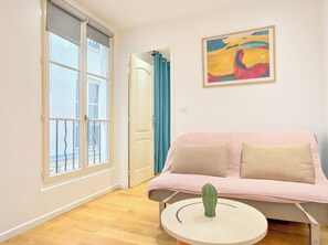 A room with a sofa, a cactus on a table, a painting on the wall, and a window with a view. - New Listing (Paris)