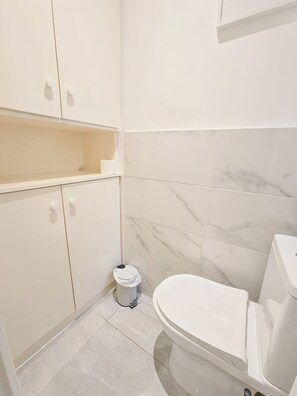 A bathroom with a toilet, a trash bin, and a cabinet with a shelf. - New Listing (Paris)