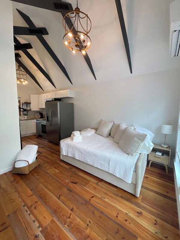 A modern bedroom with a large bed, a wooden floor, a kitchen in the background, and a unique ceiling design.