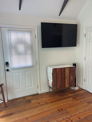 A room with a wooden floor, a white door with a window, a flat-screen TV mounted on the wall, and a cabinet with a patterned cover.