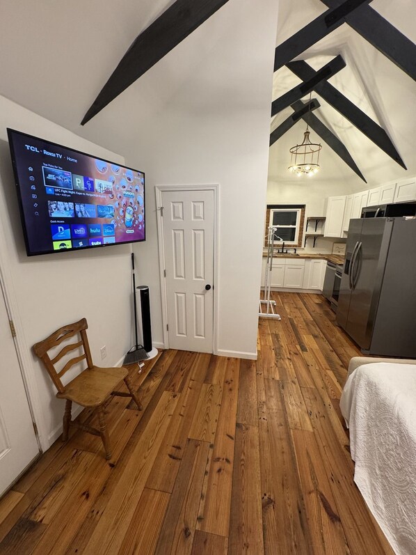 A room with a wooden floor, a white door, a television mounted on the wall, and a kitchen area visible through an open door.