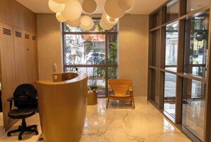 A reception area with a curved desk, a black office chair, and a wooden chair. There are potted plants and a large window with a view of the street. - New Listing (São Paulo)