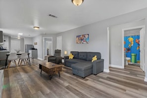 A modern living room with a grey sectional sofa, a wooden coffee table, and a dining area with chairs. - Super Mario Beach Escape (Biloxi)