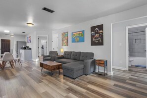 A modern living room with a sectional sofa, a coffee table, and a small side table. There is a dining area with chairs and a kitchen in the background. - New Keesler/ Beach Walking TMNT Arcade Game (Biloxi)