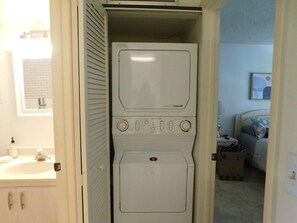 A laundry room with stacked washers and dryers, a sink with soap dispenser, and a bed in the adjacent room. - Welcoming 1-bedroom condo in sunny Sarasota with lake views! (Sarasota)