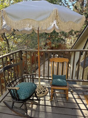 A wooden deck with a rocking chair and a chair with a cushion, a wooden table, and a striped umbrella. - Le Miel Cottage Romantic 1923 Forest Escape (Lake Arrowhead)