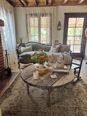 A cozy room with a vintage sofa, a round wooden table with a vase of flowers, and a bookshelf with books. - Le Miel Cottage Romantic 1923 Forest Escape (Lake Arrowhead)