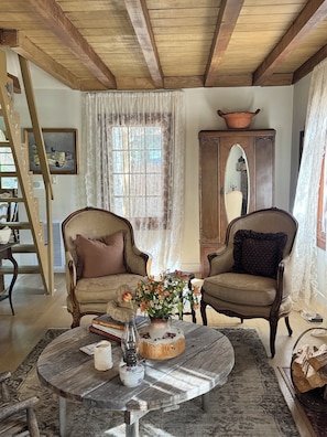 A room with two armchairs, a round table with a vase of flowers, a mirror, and a wooden ceiling. - Le Miel Cottage Romantic 1923 Forest Escape (Lake Arrowhead)