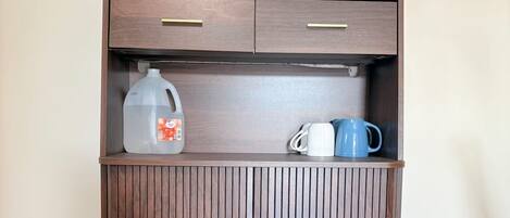 A wooden cabinet with drawers and a shelf, containing a coffee maker, a milk jug, and two mugs.
