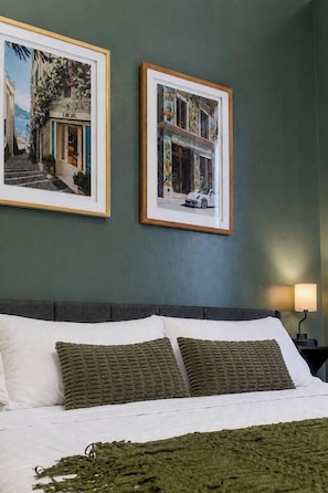 Ein Bett mit weißen Bettwäsche und grünen Kissen, ein gerahmtes Bild eines Dior-Geschäfts und ein gerahmtes Bild eines Louis Vuitton-Geschäfts an einer grünen Wand.