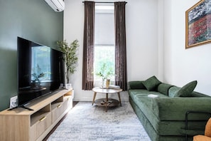 A living room with a flat-screen TV, a green sofa, a small round table with a plant, and a window with curtains. - By Yale | Sleeps 6 | Sauna + Gym | Free Parking (New Haven)