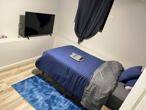 Ein Schlafzimmer mit einem Einzelbett, einem flachbildfernseher an der Wand, einer grauen Decke und einem blauen Teppich.