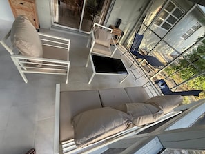 A modern outdoor seating area with white metal chairs and cushions, a small table, and a view of the surrounding landscape through large glass doors. - Bristo At Lantana (Lantana)