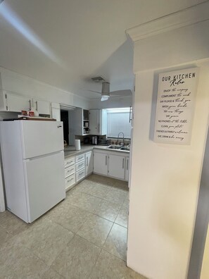 A kitchen with white cabinets, a refrigerator, a sink, and a ceiling fan. There's a sign with kitchen rules on the wall. - Bristo At Lantana (Lantana)