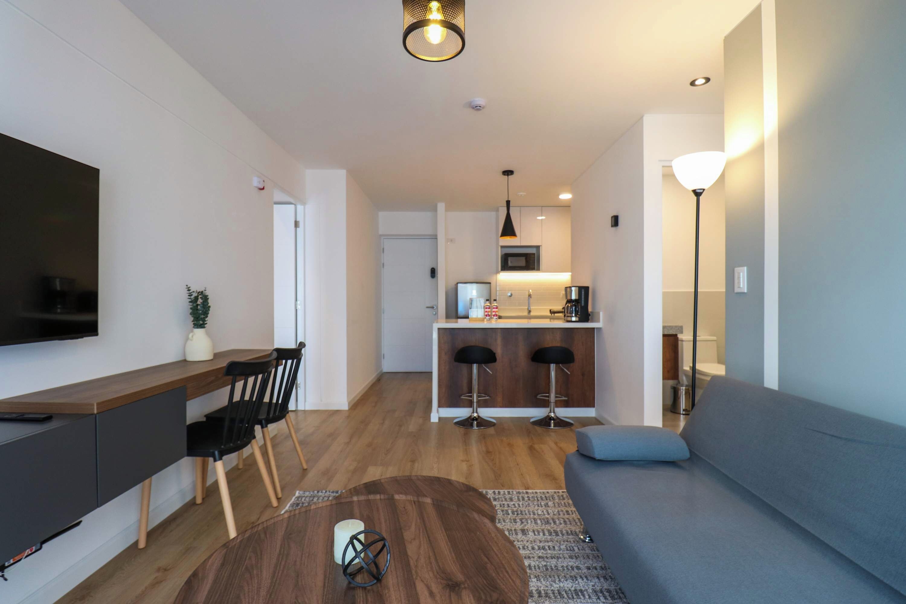 Un salon moderne avec un canapé, une table basse et un téléviseur. Il y a également un coin cuisine équipé d’un réfrigérateur, d’un four à micro-ondes et d’un évier. La pièce est dotée d’un plancher en bois et de murs blancs.