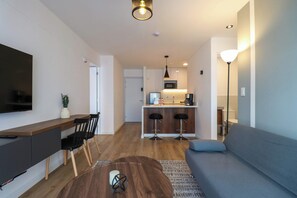 Un salon moderne avec un canapé, une table basse et un téléviseur. Il y a également un coin cuisine équipé d’un réfrigérateur, d’un four à micro-ondes et d’un évier. La pièce est dotée d’un plancher en bois et de murs blancs.