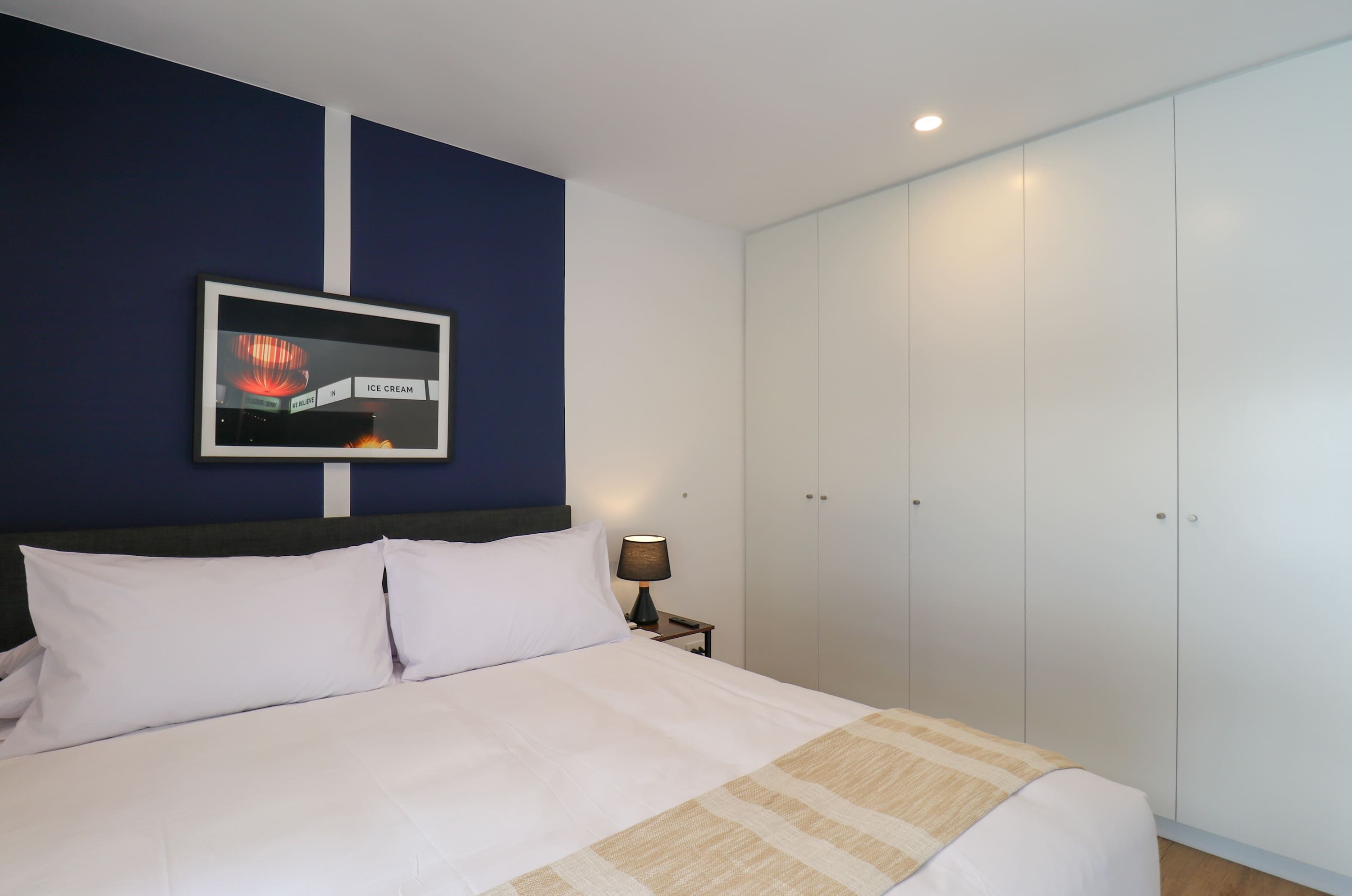Une chambre d’hôtel comprenant un lit, une table de chevet, une lampe, une armoire et un mur orné d’un tableau encadré.