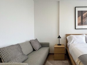 A hotel room with a grey sofa, a wooden nightstand with a lamp, a bed with white bedding, and a framed picture on the wall. - w* | Stylish Loft in the Heart of Chicó Norte (Bogotá)
