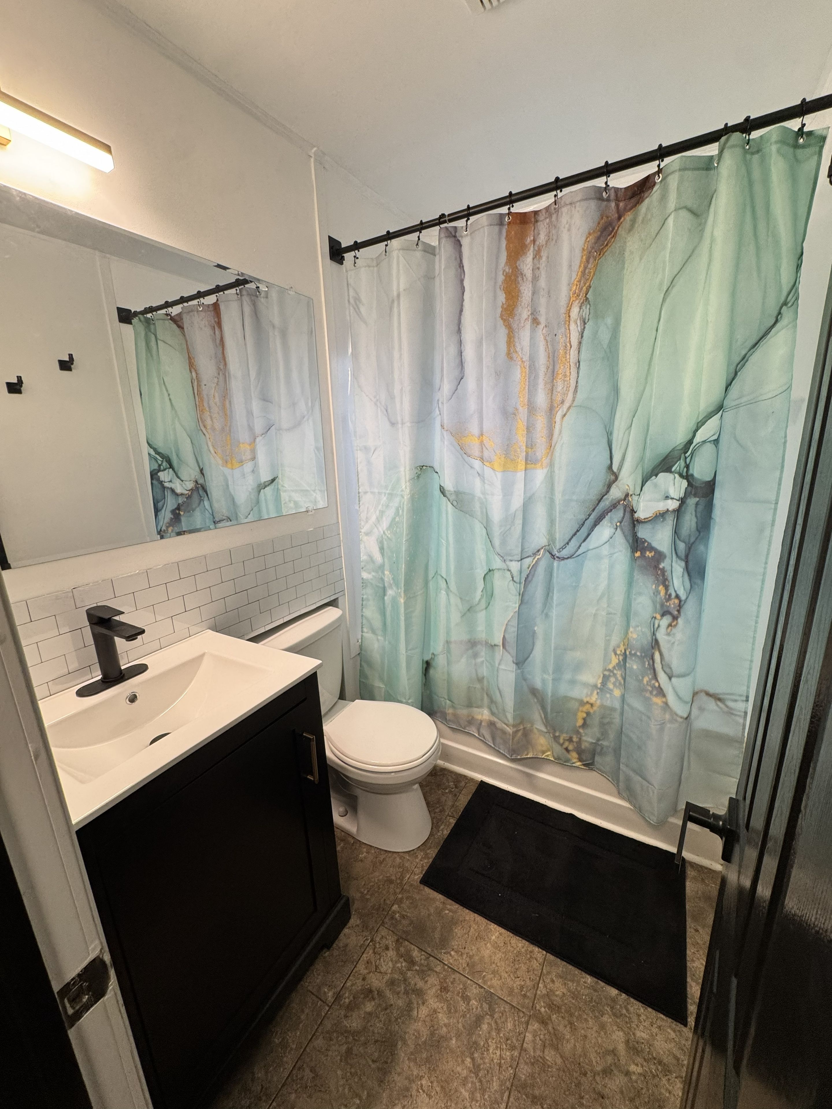 Un baño con lavamanos blanco, inodoro, cortina de ducha con diseño de mármol y un tapete negro en el suelo.