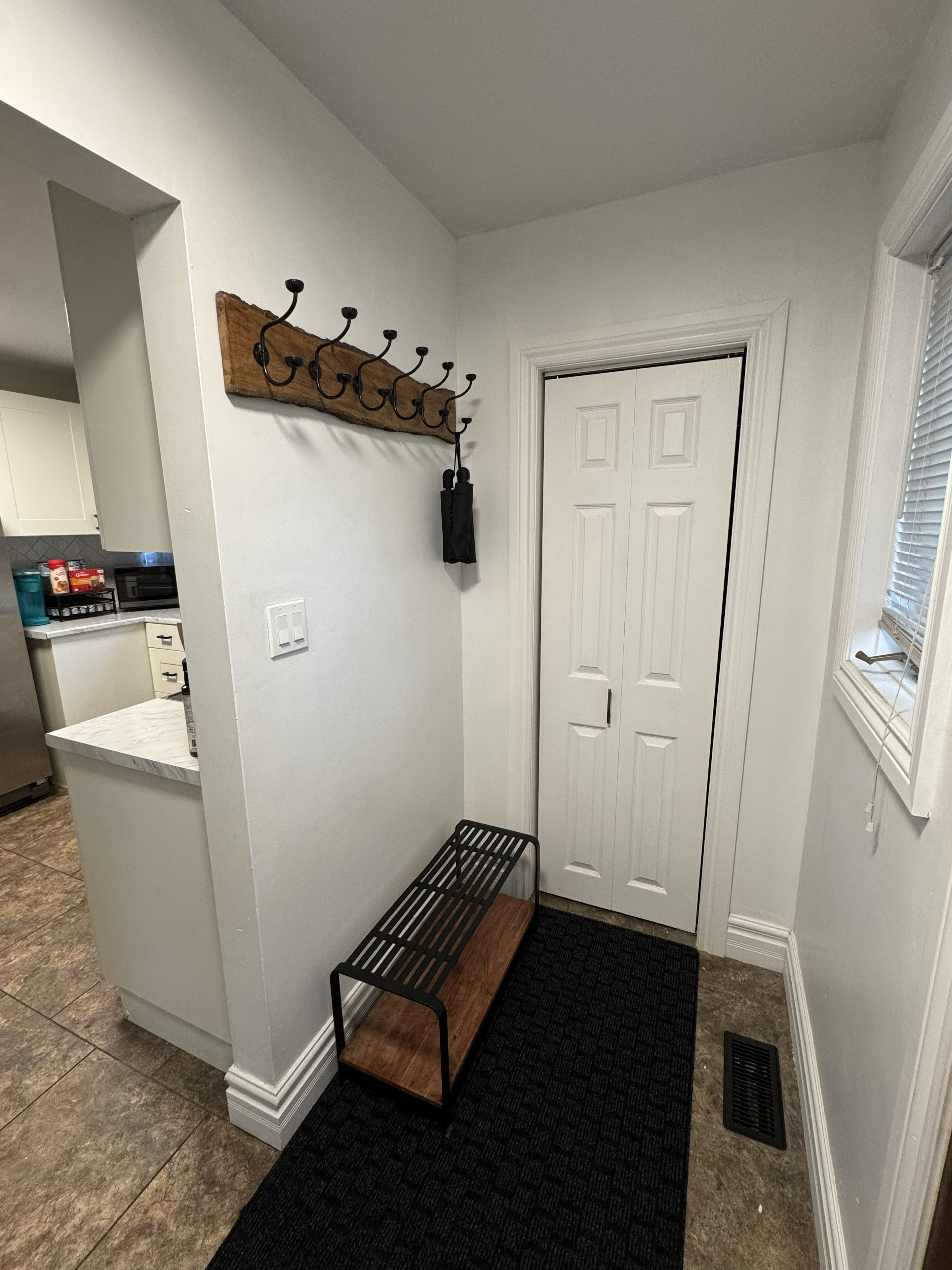 A hallway with a coat rack, a bench, a door, and a kitchen area in the background.