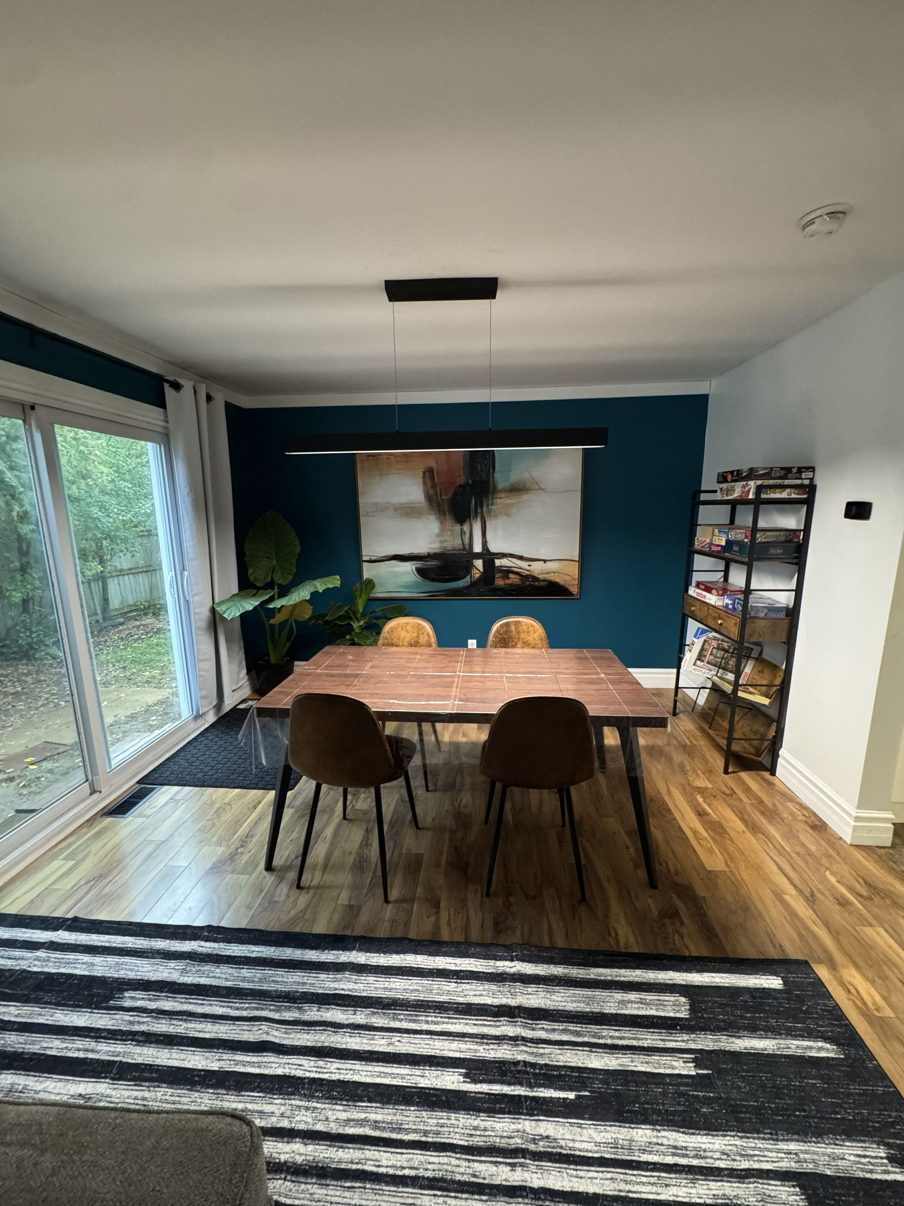 A dining area with a table, chairs, a large abstract painting, a plant, and a bookshelf.