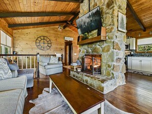 A cozy living room with a stone fireplace, wooden ceiling, and a large clock on the wall. - Cozy Cabin & Guest House Hot Tub, Firepit, Games sleeps 12 (Towns County)