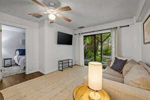 Ein modernes Wohnzimmer mit Deckenventilator, Flachbildfernseher, einer Couch, einem Couchtisch und Blick auf einen Garten durch große Fenster.