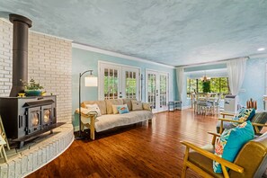 A cozy living room with a fireplace, a sofa, a dining area, and a view of greenery outside. - The Koi Cottage: Hot Tub - Treehouse - BIG Pond. 5 years as a top rated retreat! (Maggie Valley)