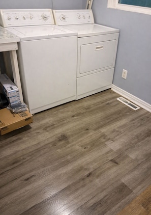 A laundry room with a front-loading washer and dryer, a vent on the floor, and a box of detergent on the floor. - Neal Street Retreat - Welcoming Home, Perfect for Families and Getaways (Tuskegee)